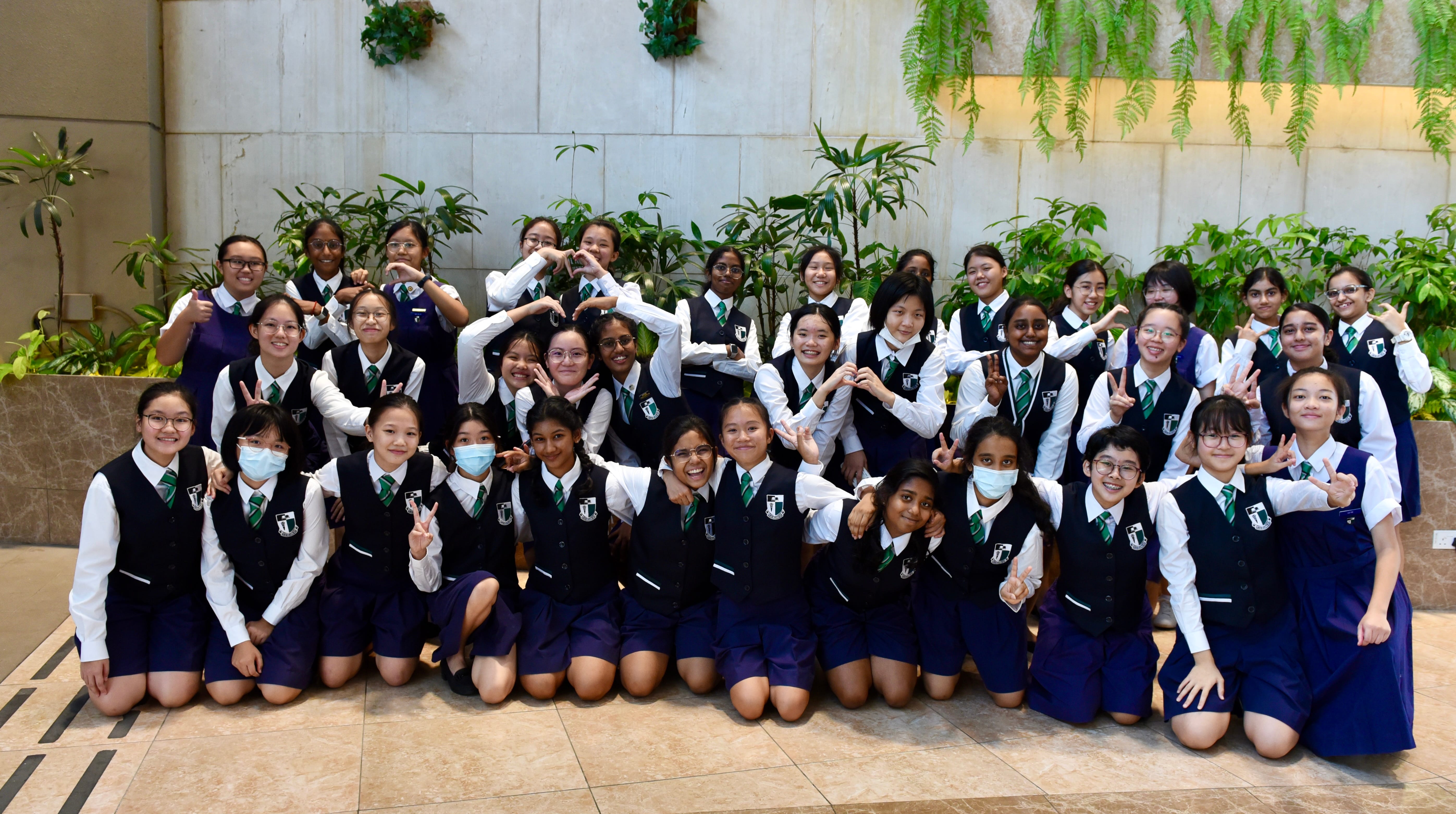 A large group of schoolgirls in navy and white uniforms with green ties pose together, making peace and heart signs.